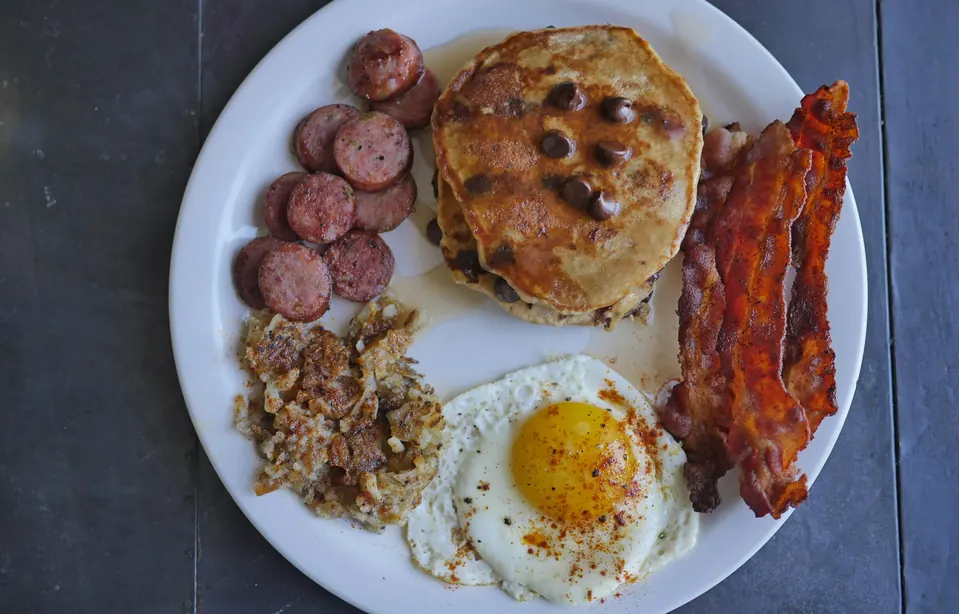 A plate of chocolate chip pancakes, bacon, sausage, hash browns and a fried egg