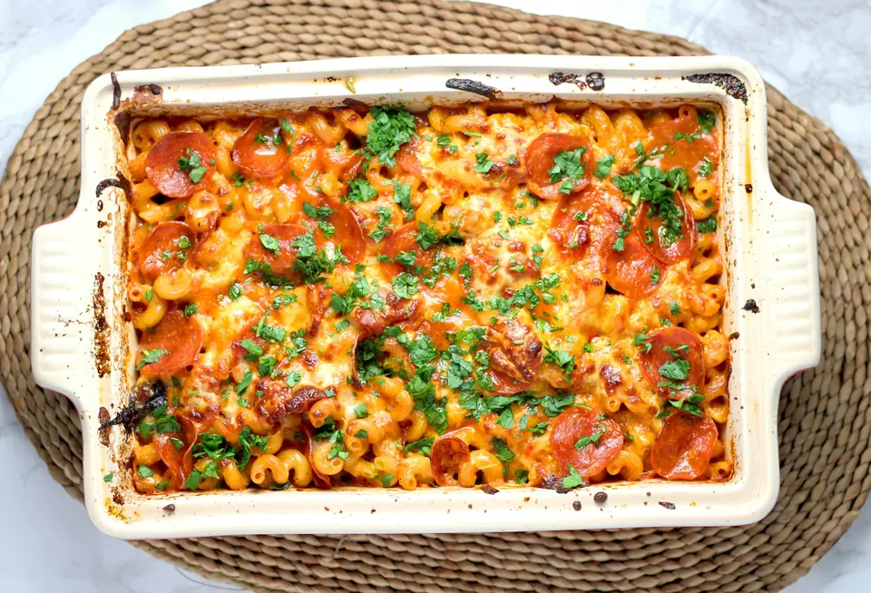 A baked pasta dish with melted cheese and herbs in a cream baking dish on a woven mat