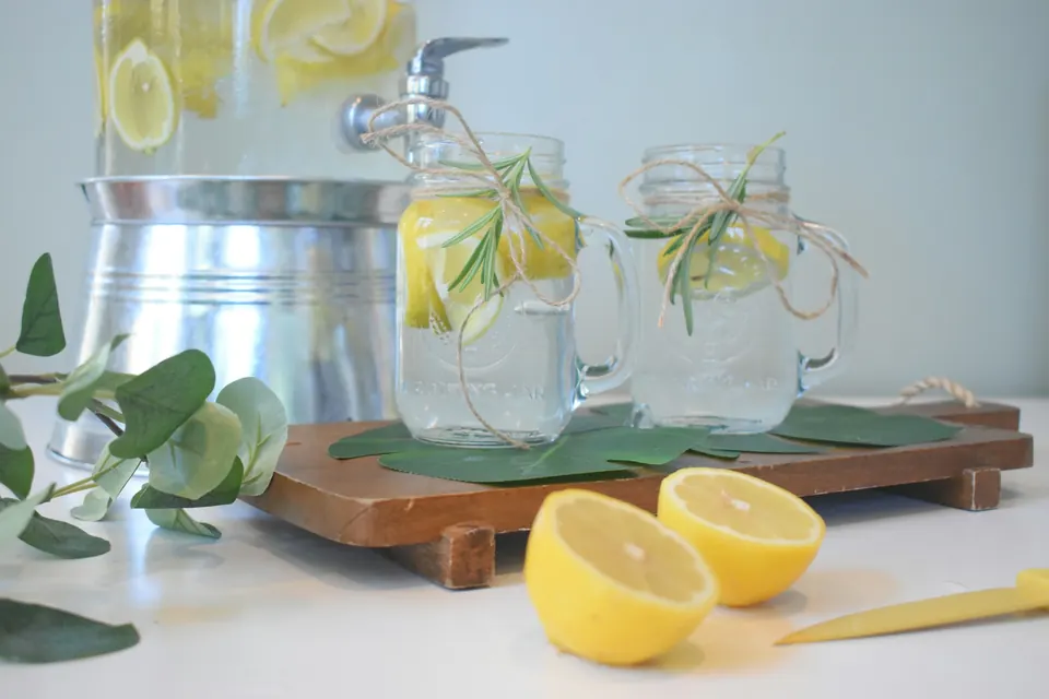 Glass jars of lemonade with rosemary beside a drink dispenser and cut lemons