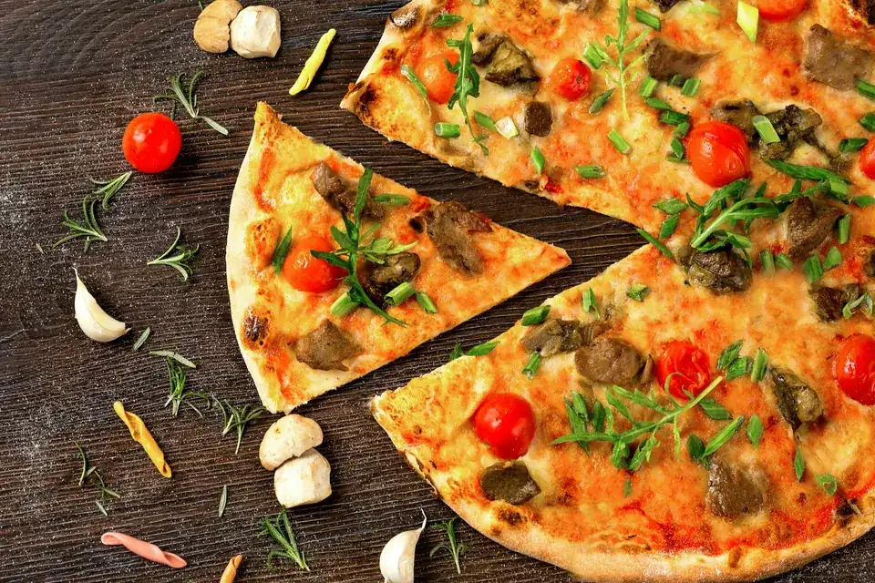 A sliced homemade pizza with tomatoes, mushrooms and herbs surrounded by scattered toppings