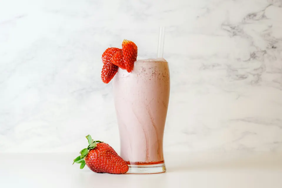 A strawberry milkshake in a tall glass with fresh strawberries on a marble counter