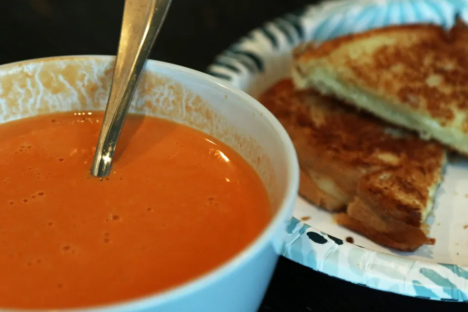 A bowl of tomato soup with a spoon beside a grilled cheese sandwich