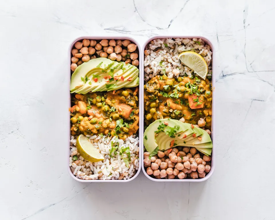Two meal-prep containers of chickpeas, rice, curry and avocado on a marble counter