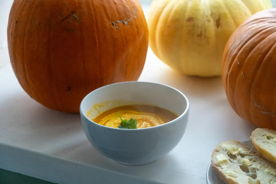 A bowl of creamy orange soup with a herb garnish in front of large pumpkins and a piece of bread