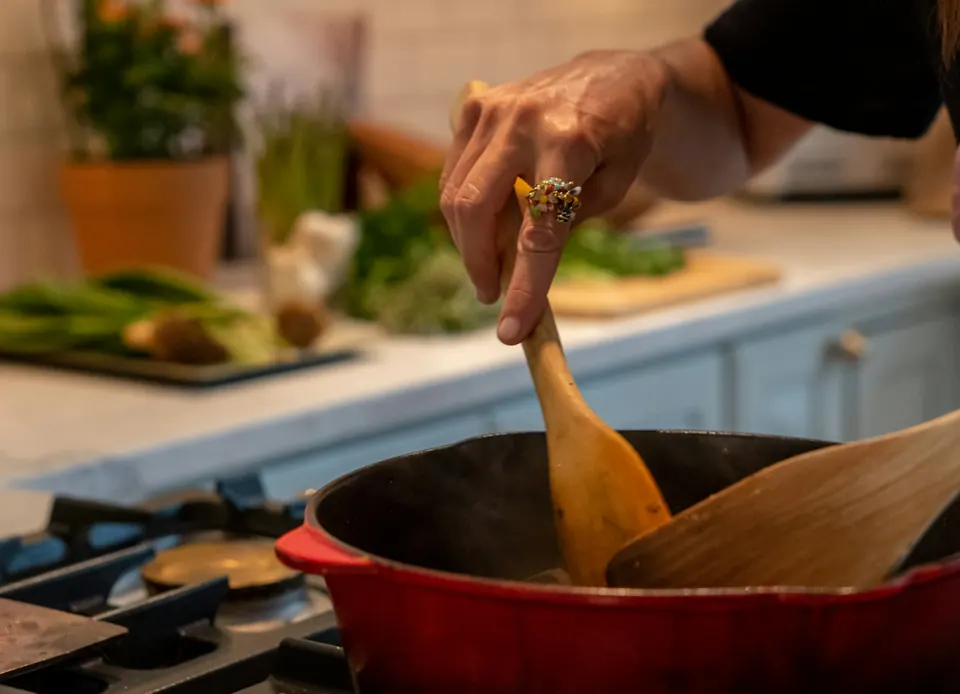 A cook stirring a red cast-iron pot with a wooden spoon at the stove