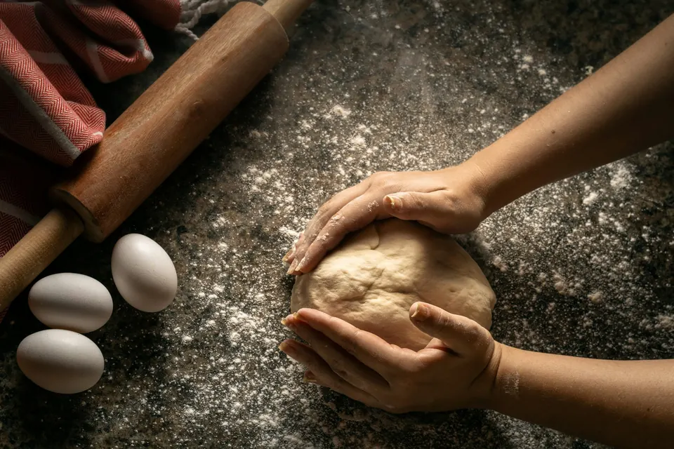Hands shaping a ball of dough on a floured counter beside a rolling pin and eggs