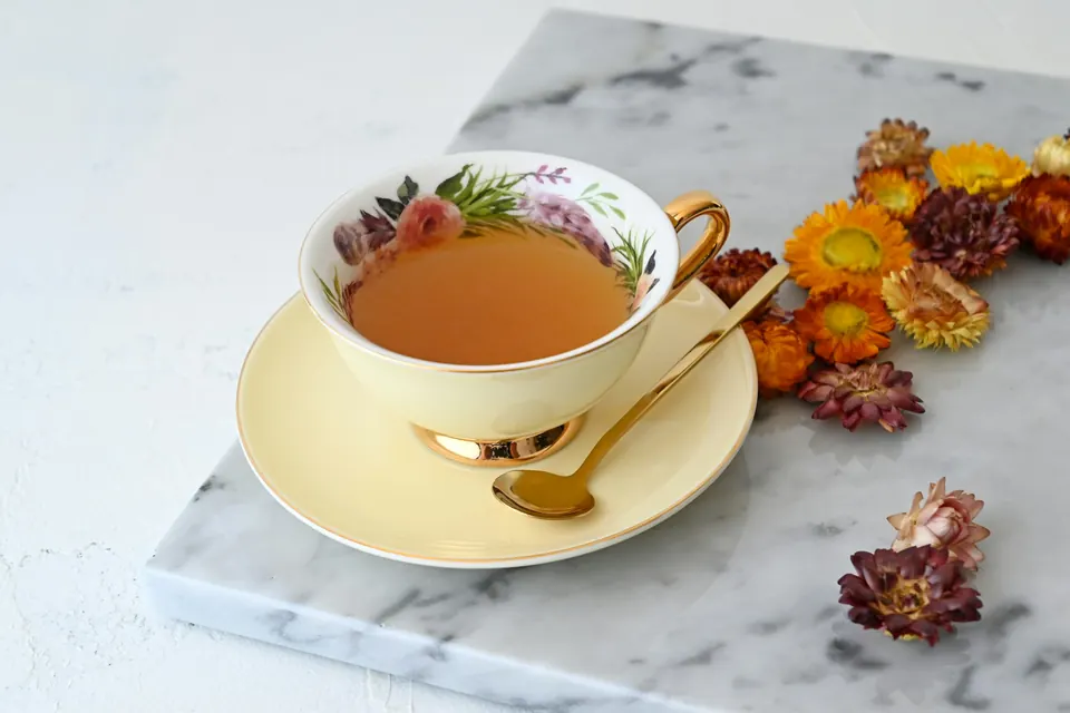A cup of tea on a saucer with a gold spoon and dried flowers on marble